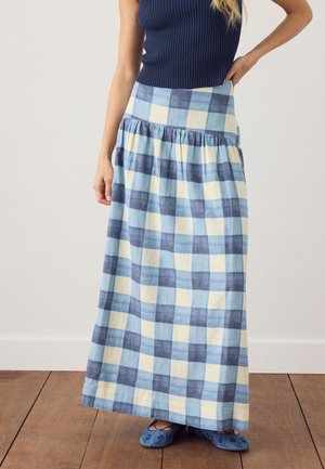 Femme portant une longue jupe à carreaux bleue et blanche associée à un top côtelé sans manches bleu marine et des ballerines bleues brodées.