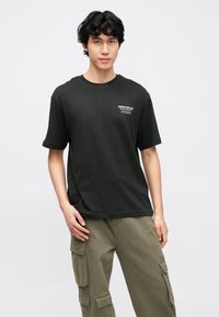 T-shirt de algodão preta de mangas curtas; apresenta um pequeno logótipo de texto branco no lado superior esquerdo; usada com calças cargo oliva.