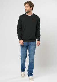 Zwarte sweatshirt met lange mouwen en een ronde halslijn, voorzien van een klein rood logo op de borst. Draag met blauwe jeans en lichte sneakers.
