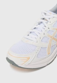 Hvit atletisk sko med mesh-overdel og oransje detaljer, plastbelægninger og gummisål. Snøring i konventionell stil.