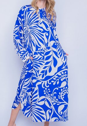 Frau trägt ein langes blau-weißes geknöpftes Kleid mit großen abstrakten Blumen- und Blattmustern und Taschen, steht vor einem einfarbigen Hintergrund.