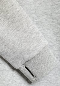 Gros plan sur une manche de sweat-shirt gris clair avec un trou pour le pouce au niveau du poignet, montrant la texture douce du tissu en maille.
