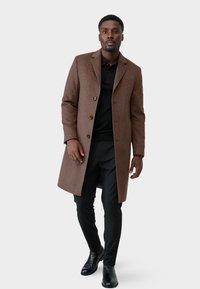 Bruine wollen overjas met een getailleerde pasvorm, enkelknoops ontwerp en drie knopen, gedragen over een zwarte polo en zwarte broek.