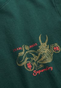 Donkergroene sweatshirt met een goud- en roodkleurig grafisch ontwerp van een mythisch wezen met horens en gestileerde tekst met de woorden "Trade Mark" en "Superdry."
