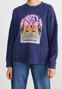 Dunkelblaues Langarm-Sweatshirt aus strukturiertem Stoff, mit einem bunten Grafikdesign, das den Text "ROXY" und "SURFING SINCE 1990" zeigt.