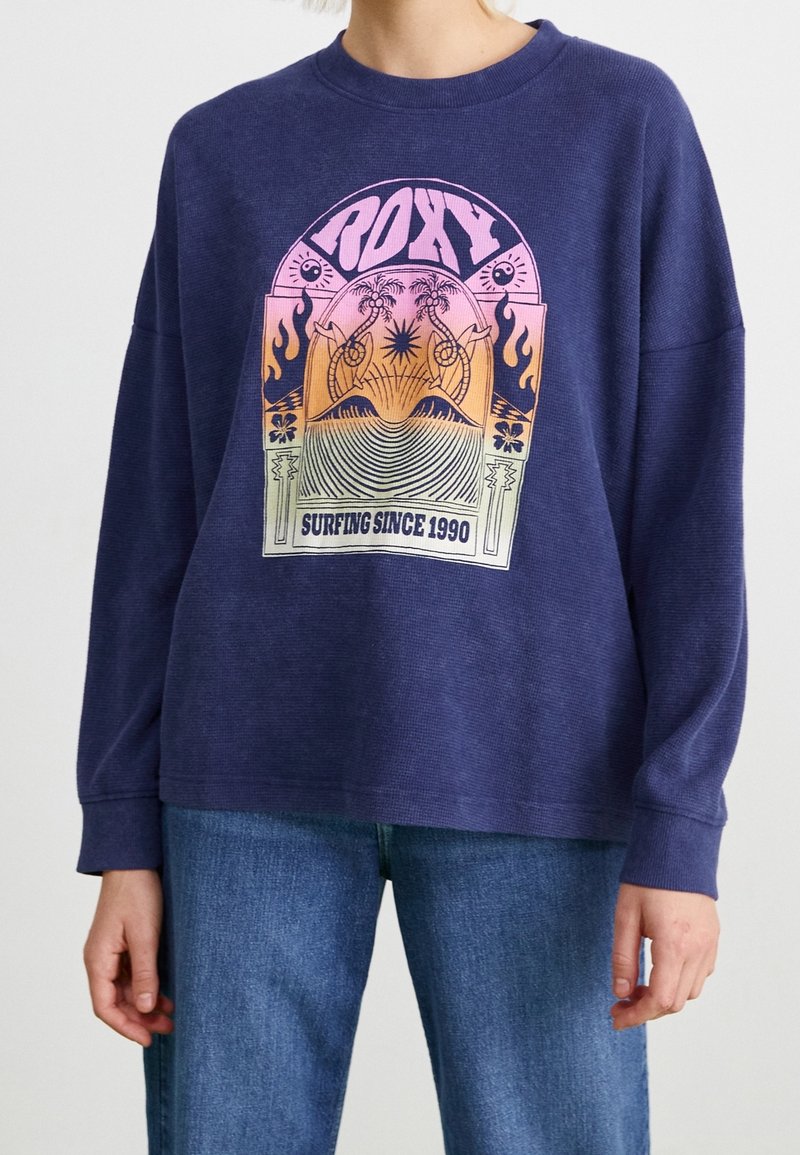 Dunkelblaues Langarm-Sweatshirt aus strukturiertem Stoff, mit einem bunten Grafikdesign, das den Text "ROXY" und "SURFING SINCE 1990" zeigt.