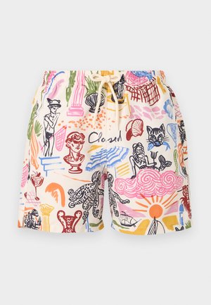 Jeune homme debout portant une chemise beige à manches longues, un short coloré à motifs et des tongs noires sur un fond blanc uni.