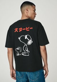 Sort bomulds T-shirt med en hvid kontur grafik af Snoopy og Woodstock på ryggen, med rød japansk tekst over designet.