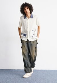 Maglietta beige a maniche corte con colletto blu navy, stampe grafiche sulla parte anteriore; abbinata a jeans blu chiaro larghi e sneakers bianche con zeppa.