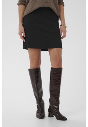 Vrouw met een zwarte minirok, bruine gestructureerde kniehoge laarzen, een bruine trui en een gouden armband aan haar rechterpols.