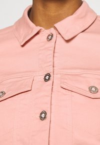 Chaqueta vaquera rosa con cuello clásico, botones plateados y dos bolsillos en el pecho con solapas. Textura suave y corte ajustado.