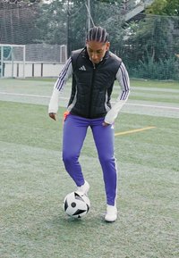 Černá prošívaná bezrukávová bunda s bílými rukávy, fialové sportovní kalhoty a bílé tenisky. U nohy leží černobílý fotbal.