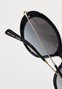 Ochelari de soare ovali negri cu lentile gri gradient, având detalii din metal auriu și brațe negre gravate cu "MICHAEL KORS."