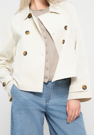Frau trägt eine cremefarbene, kurze, zweireihige Jacke mit Schildpattknöpfen, eine beige, geknöpfte Strickjacke und blaue Jeans.