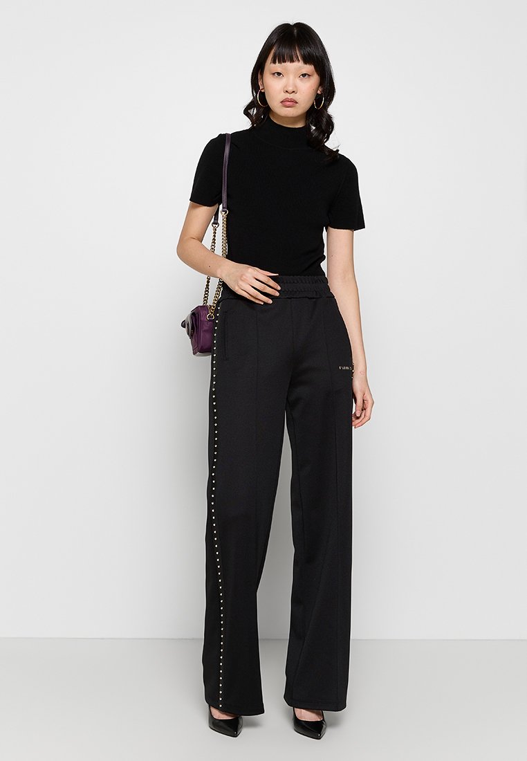 Donna che indossa un dolcevita nero a maniche corte, pantaloni neri larghi con borchie laterali, tacchi neri, orecchini a cerchio dorati e che porta una borsa a tracolla viola.