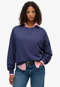 Granatowy sweatshirt z ryflowanym wzorem i luźnym krojem, nałożony na jasnoróżową, w paski koszulę z guzikami. Czarny pasek ze srebrną klamrą.