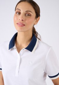 Polo blanc avec des accents marine et crème, tissu texturé, patte de boutonnage à trois boutons et petit logo sur la poitrine gauche.