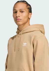 Bež kapuca s kengursko žepom, kapuljača sa špagom i bijelim Adidas logotipom na prsima. Mekana tkanina s glatkom teksturom.