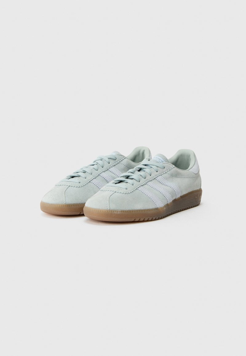 Par de zapatillas deportivas de ante gris claro con suelas de goma color caramelo y detalles de tres franjas blancas, mostradas sobre un fondo blanco liso.