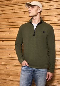 Olivově zelený svetr na zip s texturovanou látkou, černým zipem a malým černým logem. Slučuje se se světle modrými džíny a béžovou čepicí.