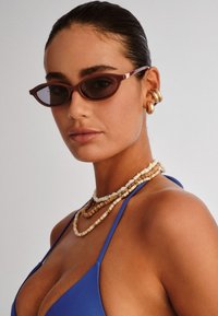 Brown cat-eye sunglasses with dark lenses, paired with layered beaded necklaces, featuring various textures and colors, against a neutral background.