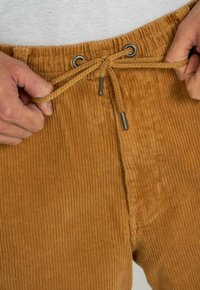 Corduroy trousers in a warm brown color with vertical ribs, featuring a drawstring waist and metal eyelets. Textured fabric detail is visible.