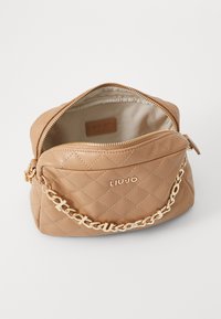 Bolso de mano acolchado tan con cadena dorada y cierre de cremallera. Presenta un parche de logo de cuero y un forro interior beige. Diseño compacto.