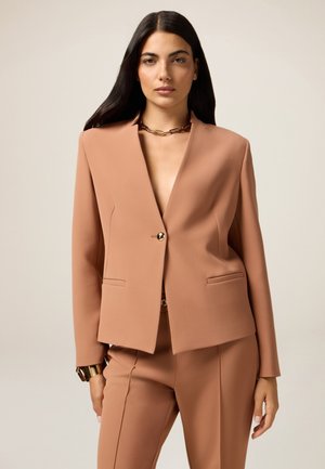 Blazer pesca con revers, chiusura a un bottone dorato e due tasche frontali. Indossato con pantaloni abbinati e una collana a catena dorata.
