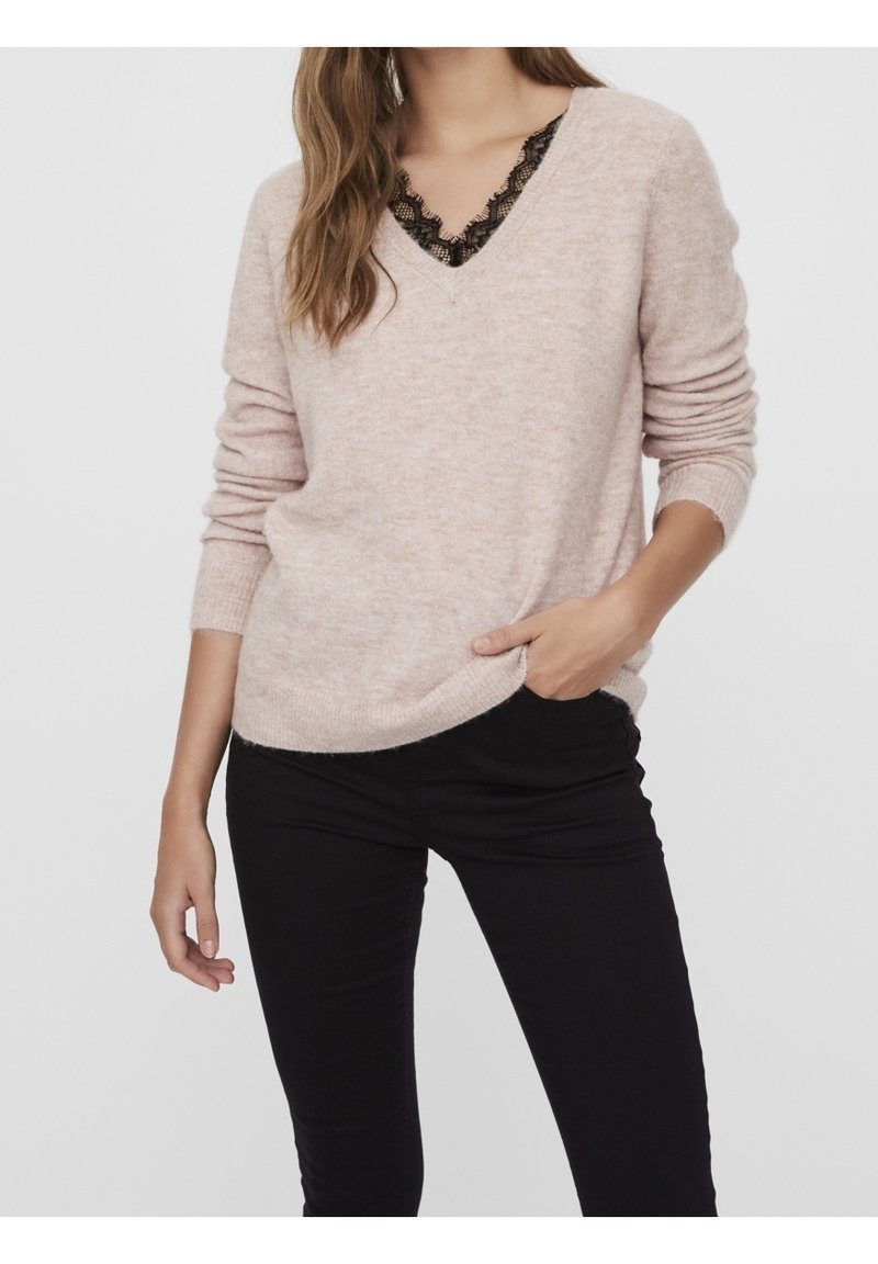 Femme portant un pull col V rose clair avec un bord en dentelle noire visible, associé à un pantalon noir ajusté, main reposant dans la poche.