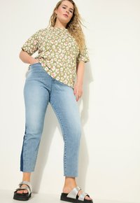 Olivově zelená košile s krátkým rukávem, zdobená růžovým a bílým květinovým vzorem, v kombinaci se světle modrými džíny s tmavě modrými postranními pruhy a černými sandály.