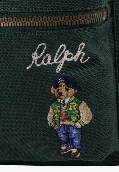 Zelena tkanina z zlatim zadrgo, izvezeno belo besedilo "Ralph" in podroben izvezen medved, oblečen v vsakodnevna oblačila in kapo.