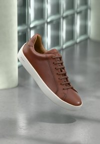 Bruine leren sneaker met witte rubberen zool, voorzien van een gladde textuur, een ronde neus en veters. Merkenlogo zichtbaar aan de zijkant.