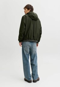 Mørkegrøn hættetrøje med elastisk kant og manchetter, kombineret med lyseblå løstsiddende jeans. Materialet virker glat og let.