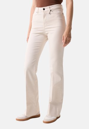 Frau trägt hoch taillierte, cremefarbene Jeans mit geradem Bein und weiße Sneakers, steht vor einem schlichten hellen Hintergrund.