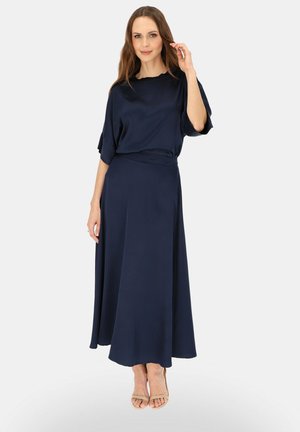 Kobieta ubrana w luźną granatową bluzkę i dopasowaną długą spódnicę, stoi z jedną ręką uniesioną blisko twarzy, beżowe buty na otwartym palcu na obcasie.