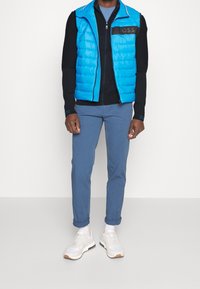 Blå puffy väst över en svart tröja, kombinerad med blå byxor och vita sneakers. Västen har en svart logotyp på bröstet.