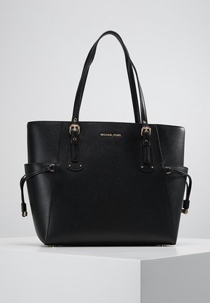 Sac cabas noir Michael Kors avec doubles bretelles, poches latérales et détails de boucle dorée sur une surface blanche avec un fond gris.