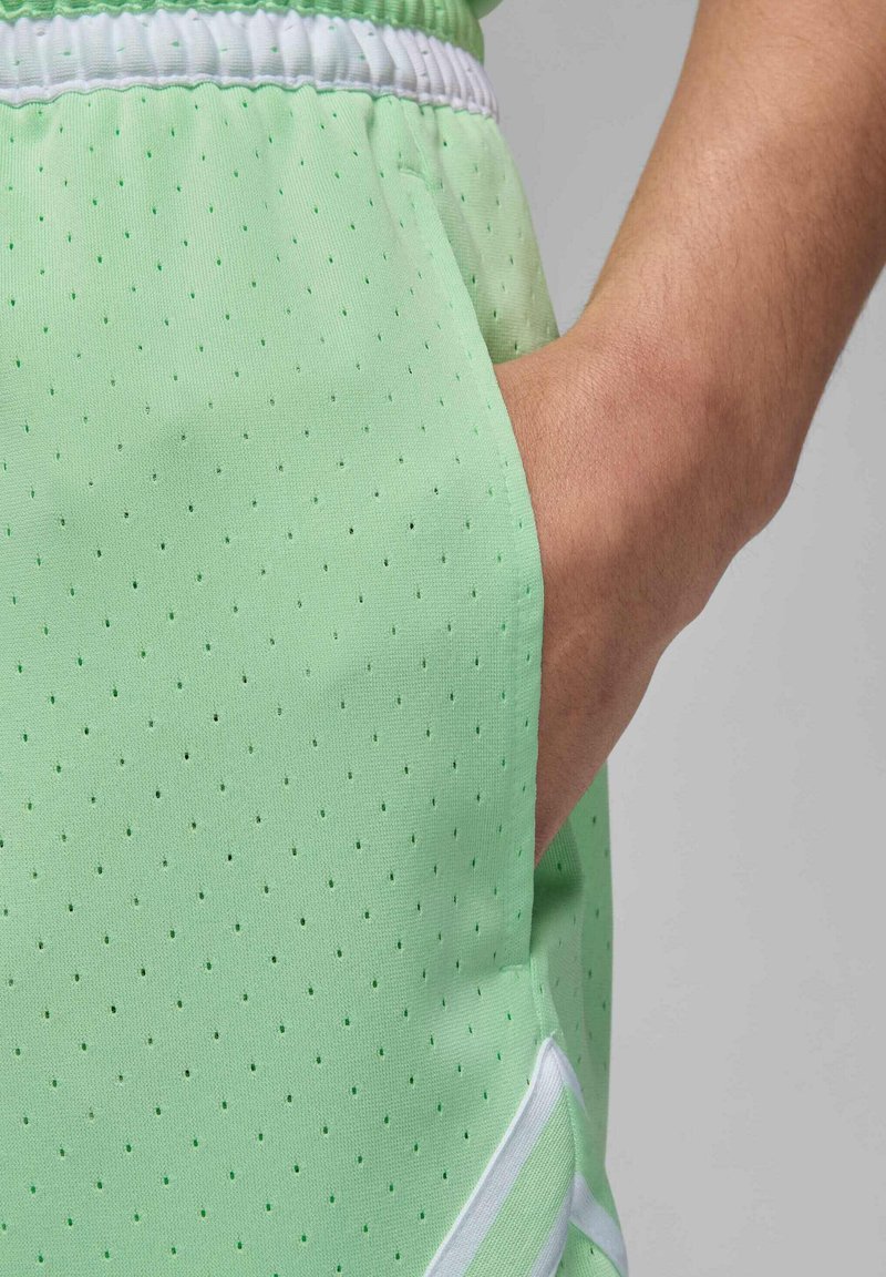 Pantalones cortos deportivos verde claro con textura perforada, que presentan una cinturilla elástica blanca y un bolsillo lateral. Los detalles incluyen un ribete blanco.