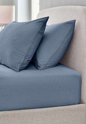 Zwei blaue Kissen und ein blaues Spannbettlaken auf einem beigen gepolsterten Bettrahmen in einem sanft beleuchteten Schlafzimmer.