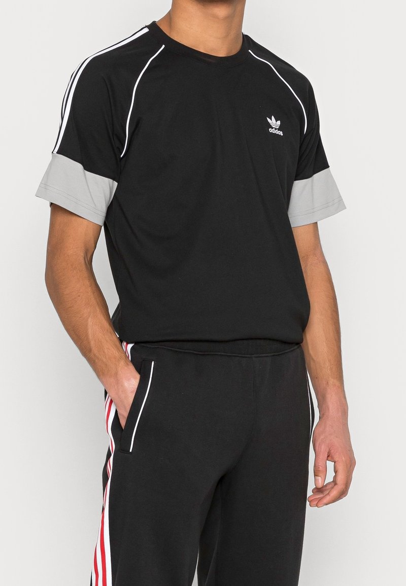 Maglietta sportiva nera con maniche corte grigie, dettagli bianchi e logo adidas sul petto, abbinata a pantaloni neri con strisce rosse.