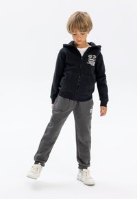 Schwarze Zip-Hoodie mit Fronttaschen und grafischem Text, kombiniert mit grauen Jogginghosen mit Seitentaschen und einem Logo-Patch. Weiße Sneaker.