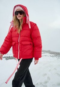Czerwona kurtka puchowa z różowymi akcentami, wyposażona w kaptur, dwie przednie kieszenie oraz sznurki ściągające na dole, noszona z czarnymi spodniami na zaśnieżonym tle.