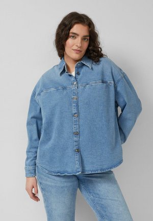 Kobieta z kręconymi brązowymi włosami, ubrana w dużą zapinaną na guziki denimową koszulę i jasnoniebieskie dżinsy, stojąca na tle jednolitego szarego tła.