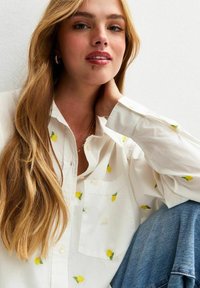 Chemise blanche à boutons avec broderie de citrons jaunes, manches longues et poche poitrine. Le mannequin a les cheveux longs et est assis contre un mur.