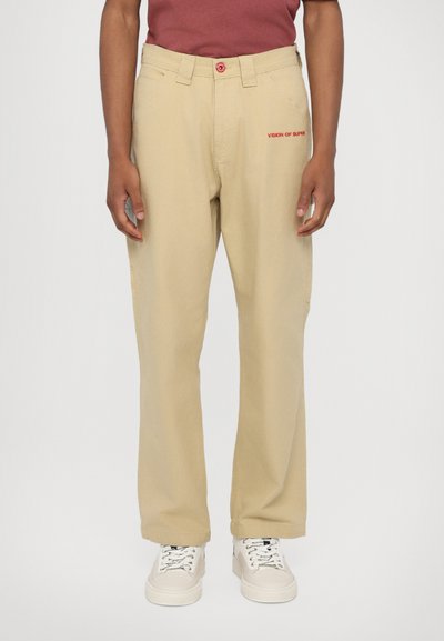 Beige cargo bukser med en afslappet pasform, som har forlommer og broderet tekst i rød. Båret med hvide sneakers.