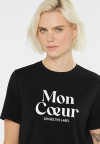 Black t-shirt featuring white text "Mon Coeur" and "SENSES.THE LABEL." Soft fabric, short sleeves, round neckline, casual fit.