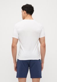 T-shirt blanc à manches courtes en tissu lisse, vu de dos. Porté avec un short bleu foncé, présentant un design simple.