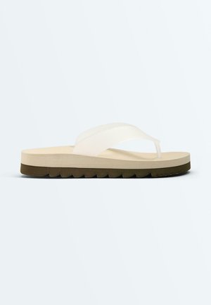 Jedna béžová žabková obuv s hrubou podrážkou a bielym remienkom, zobrazená z bočného pohľadu na jednoduchom svetlosivom pozadí.