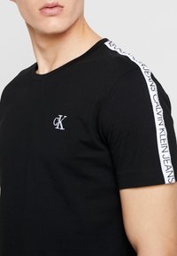 Černé bavlněné tričko s kulatým výstřihem, na kterém se nachází malé bílé vyšívané logo a kontrastní bílé boční pruhy s textem značky.