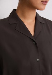 Camicia nera con bottoni, caratterizzata da un colletto rilassato, una texture liscia e un dettaglio con un solo bottone, che mostra un design minimalista.